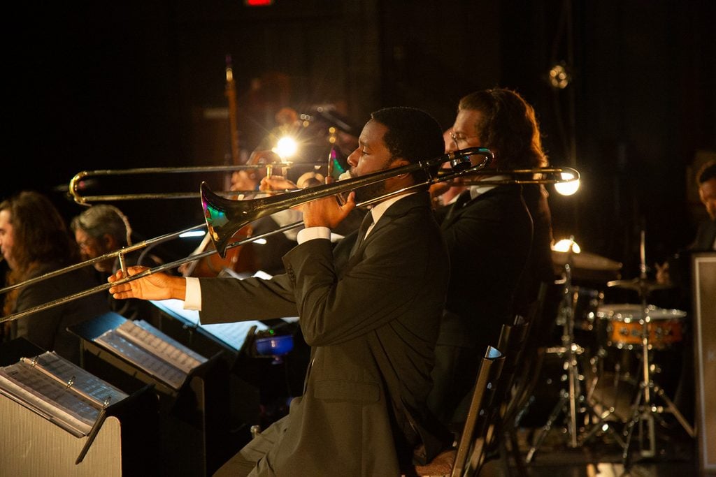 Two trombone players in black suits playing on a stage with a black background.