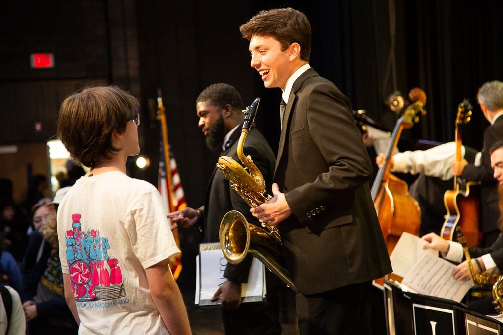 Man in a black suit, holding a baritone saxophone laughing and talking to a male student in a white t-shirt in an auditorium