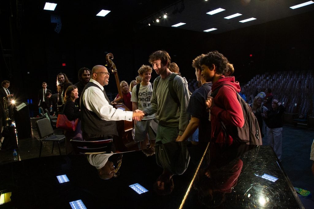 John Brown, holding his double bass shaking hands with a male high school student in a gray sweatshirt and surrounded by five other male students with the band in the background.