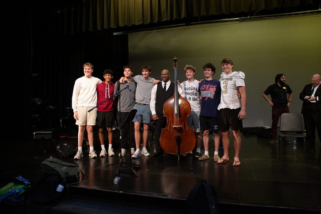 John Brown holding his double bass with 4 male high schooler to his left and 3 male high schoolers to his right, all posing for a photo.