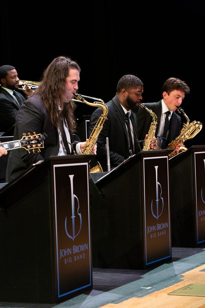 Three saxophone players in balck suits seated on stage playing. Music stands in from of them feature the John Brown Big Band logo.