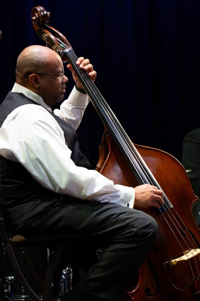 John brown in a white button down shirt, black vest, and black slacks, sitting on a chair while playing the double bass.