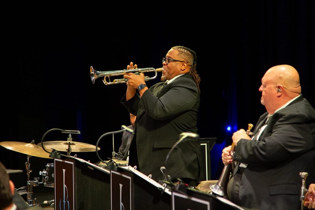 Male trumpet player in a black suit standing and playing a solo on stage with a black background. Another trumpet player sits next to him.