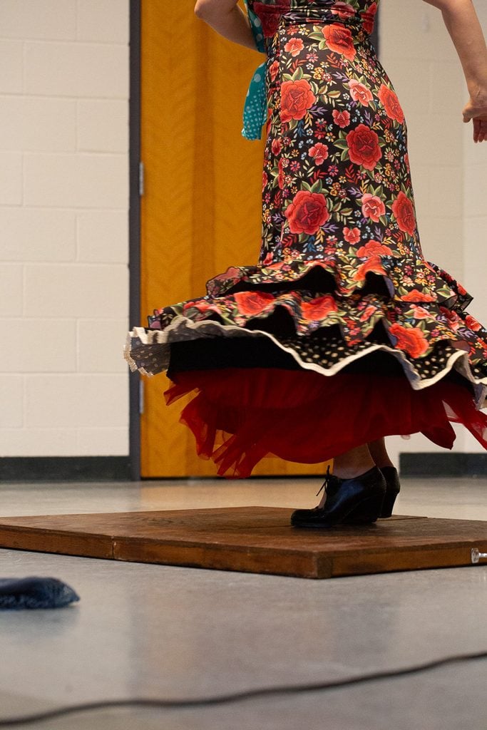 Flamenco dancer in a red floral dress twirls with her dress flaring out while her shoes dance on a wooden board.