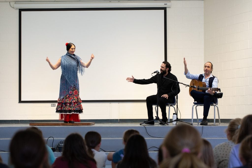A flamenco in a blue shawl and patterned red skirt gestures on a stage while two musicians play instruments—one with an acoustic guitar beside a microphone—before an audience in front of a blank projection screen.