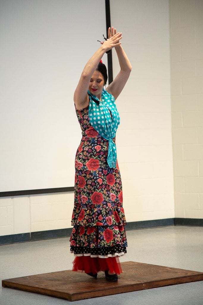 Flamenco dancer in a red floral dress with turquoise polka dot scarf dances with her arms raised above her head.