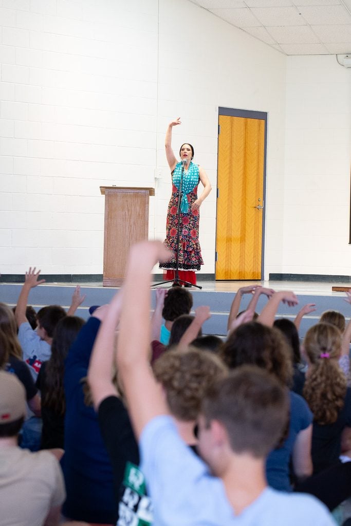 Flamenco dancer in a red floral dress with turquoise polka dot scarf stands on stage in front of students with one arm raised as students in the audience raise one arm as well in response.