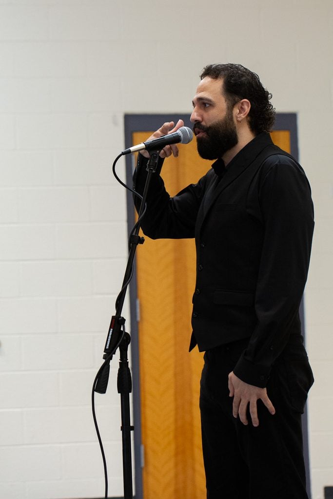 Male singer dressed in a black vest, button down shirt and slack, since on stage in front of a microphone with one hand raised near his mouth.