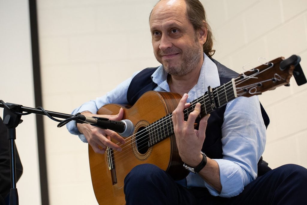 Guitarist in a black vest and light blue button down shirt smiles at audience from his seat on the stage while strumming his guitar.