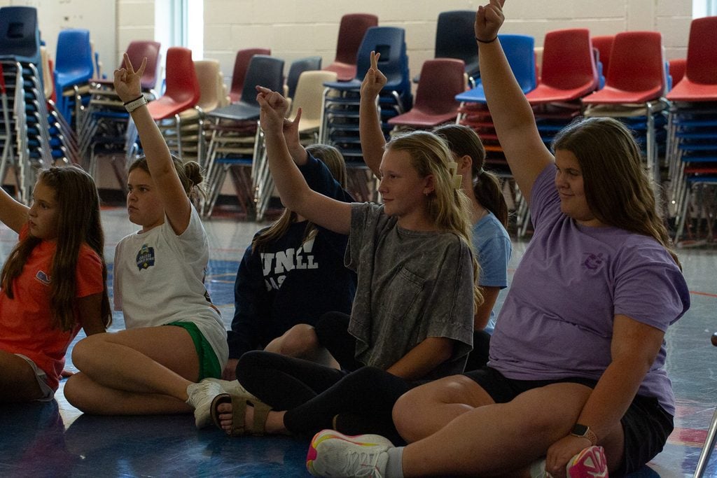 Six girls sitting on a gym floor raise their hands in the air with pinky and ring fingers extended.