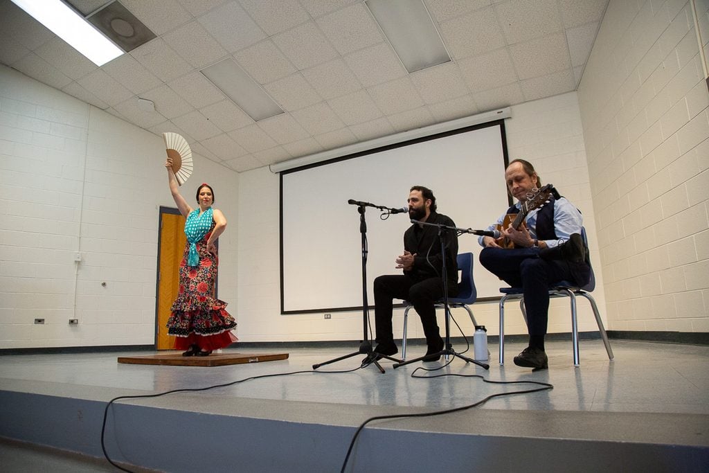 A flamenco dancer is a red floral dress with turquoise scarf holds a fan above her head while two musicians sit on stage near her - one singing in clapping, the other playing a guitar.