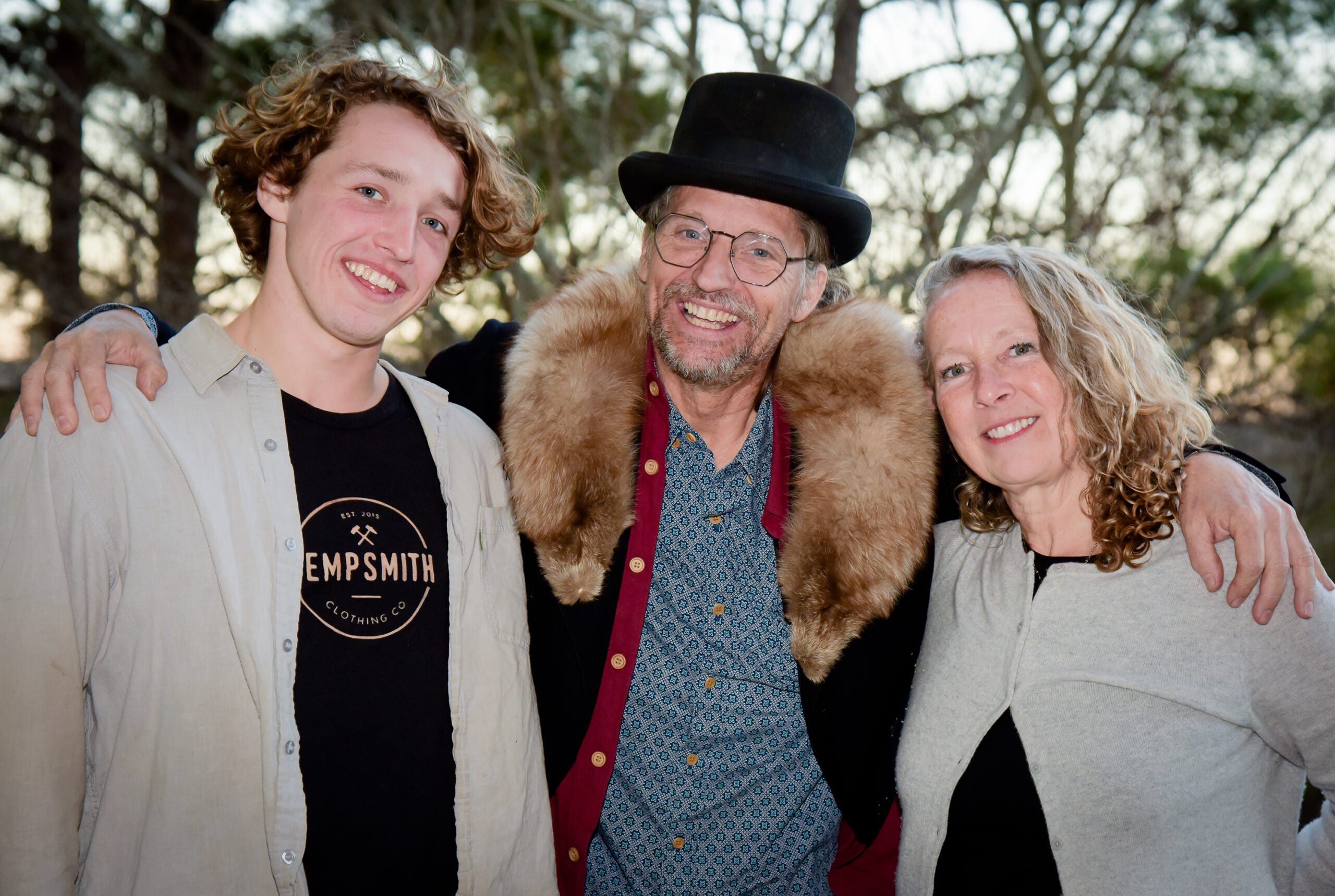 A man in a top hat, center, has his arms around a woman and a young man. They are all smiling for the camera.