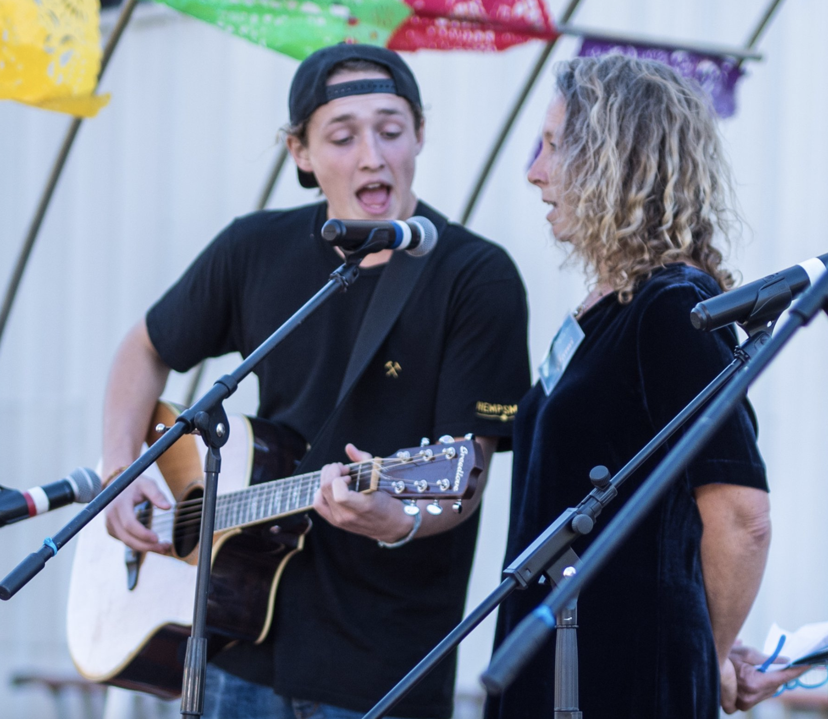 A young man on guitar wearing a black baseball cap backwards shares a microphone with a blonde woman, also wearing black. They are singing.