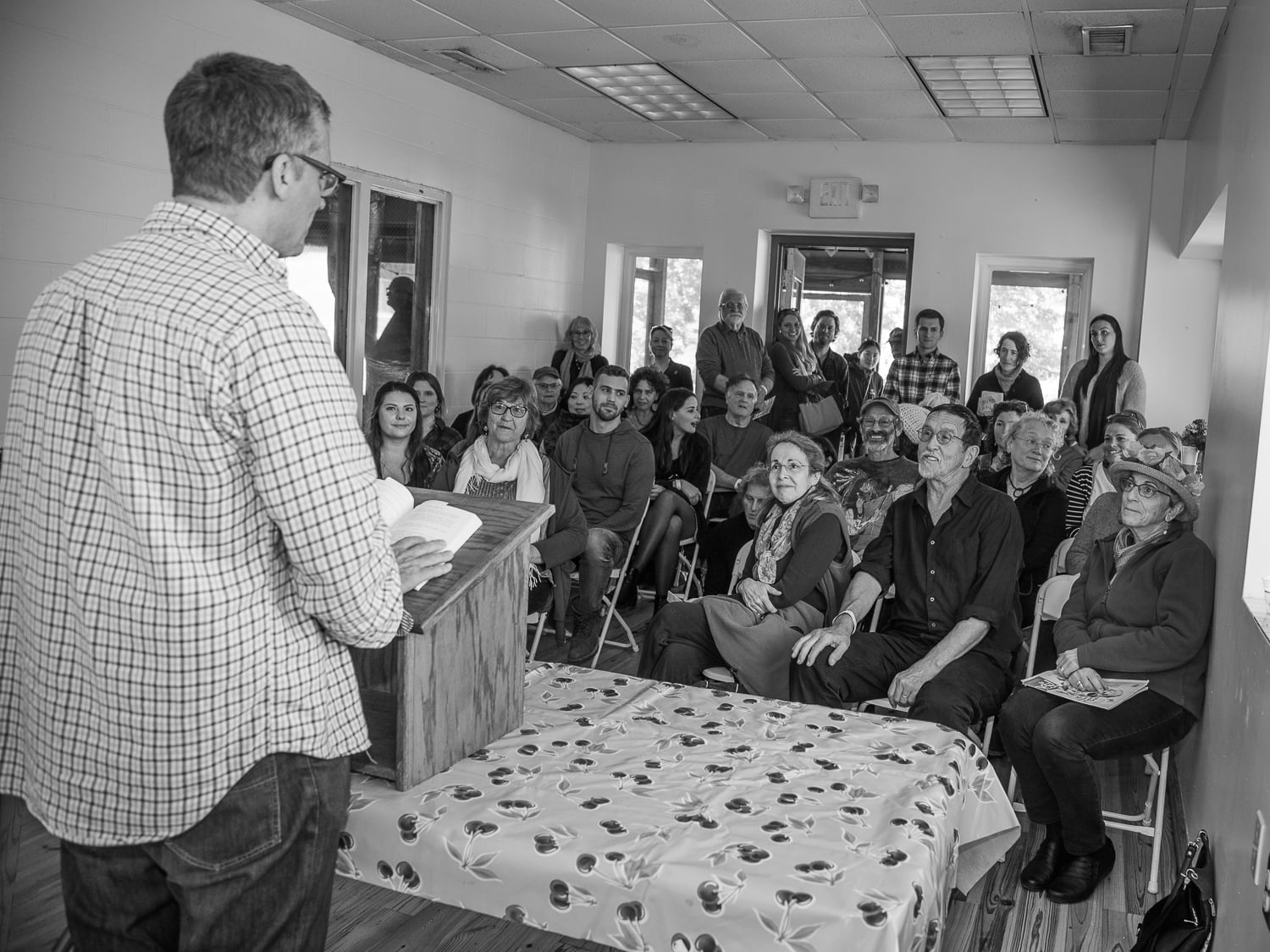 An audience of people gathered closely in a room observe a man speaking at a lectern.