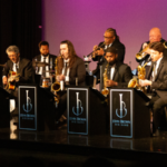 John Brown's Little Big Band playing on stage in black suits in front of a pink and teal lit background.