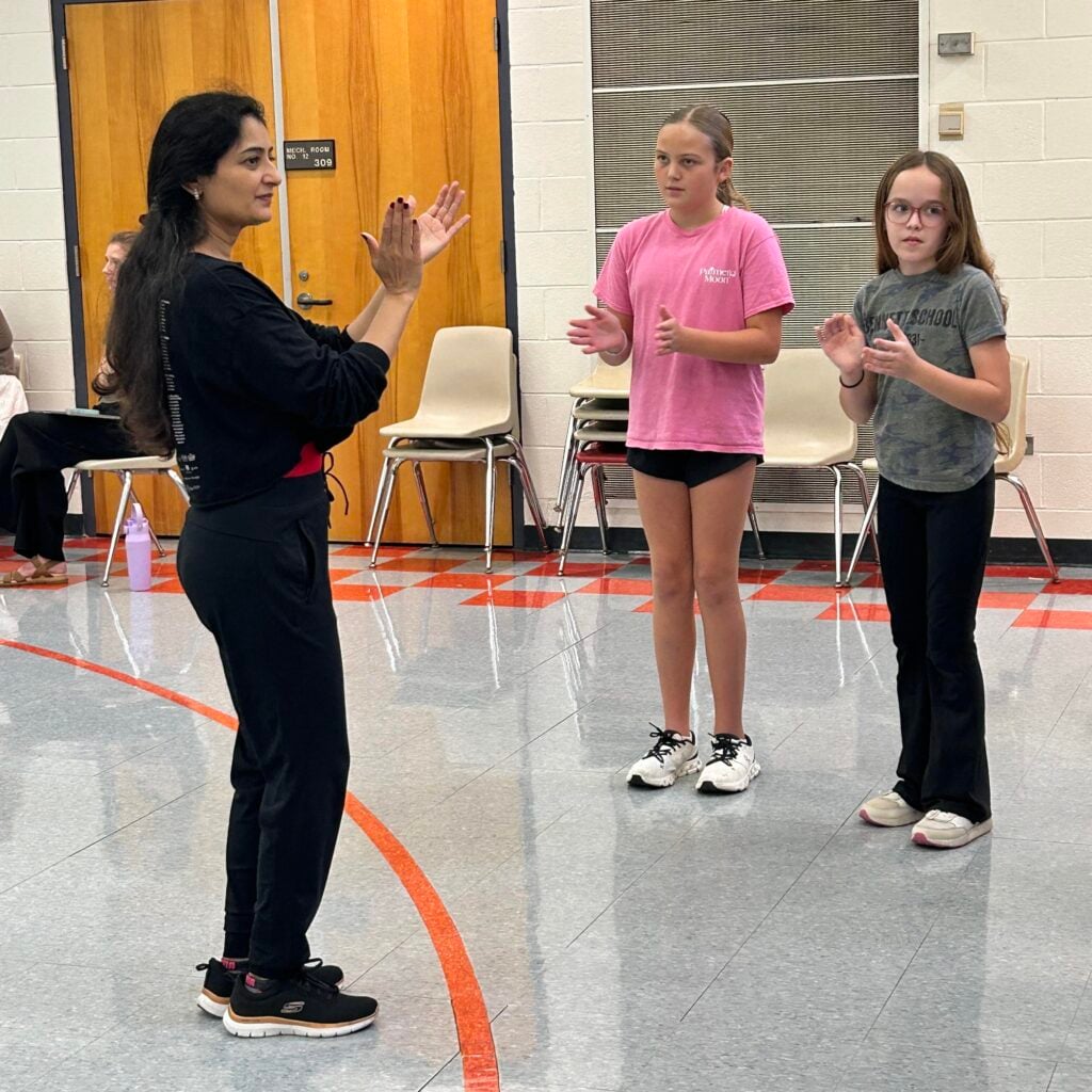 Flamenco dancer in black sweatshirt and pants, claps out a rhythm with two girls who clap with her inside a gym.
