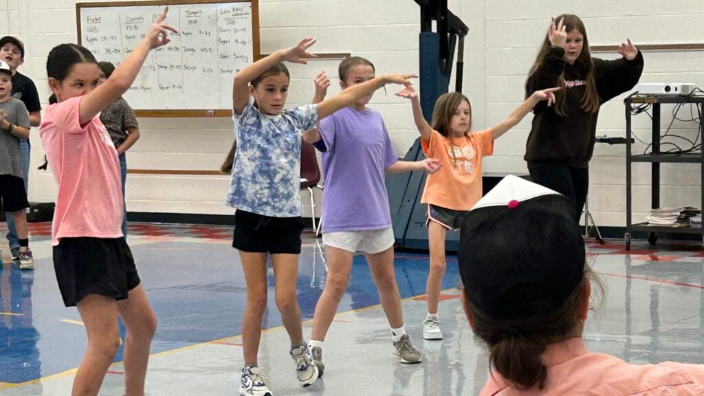 Five girls practice flamenco dance steps with their arms swinging above their heads while the guitarist plays music in front of them