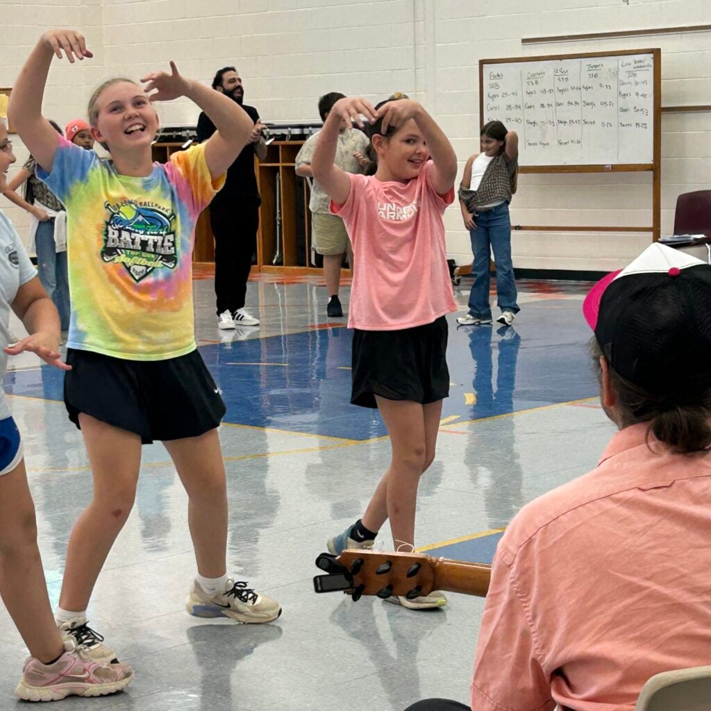 Two girls in shorts and t-shirts practice a flamenco dance move with their arms raised and smiles on their faces while the guitarist plays music in front of them